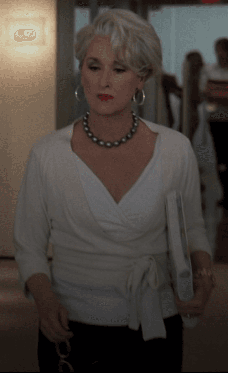 Miranda Priestly embodies effortless authority in a sophisticated cream wrap top, accessorized with a striking dark pearl necklace, navigating the office with her signature composed demeanor.