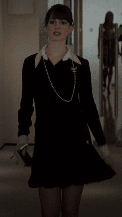 Andy Sachs navigates the demanding world of high fashion in a chic black mini dress, elegantly framed by a crisp white collar and cuffs, adorned with a timeless pearl necklace that adds a touch of classic sophistication.